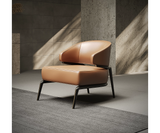 A modern armchair with a curved leather seat and backrest, supported by a sleek metal frame. The armchair is placed on a grey rug in a minimalist room with concrete walls and natural light.